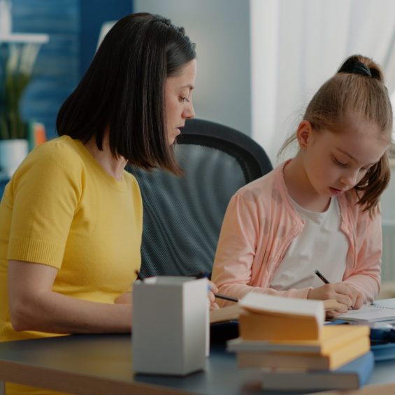 Pupil and mother doing homework together with books at desk. Young girl writing on textbook, preparing for online classes and school tasks with help from parent with supplies and book